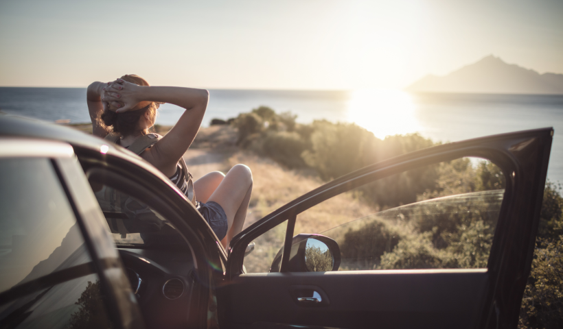 En kvinna sitter bekvämt på motorhuven på sin elbil, njuter av solen och havsutsikten.